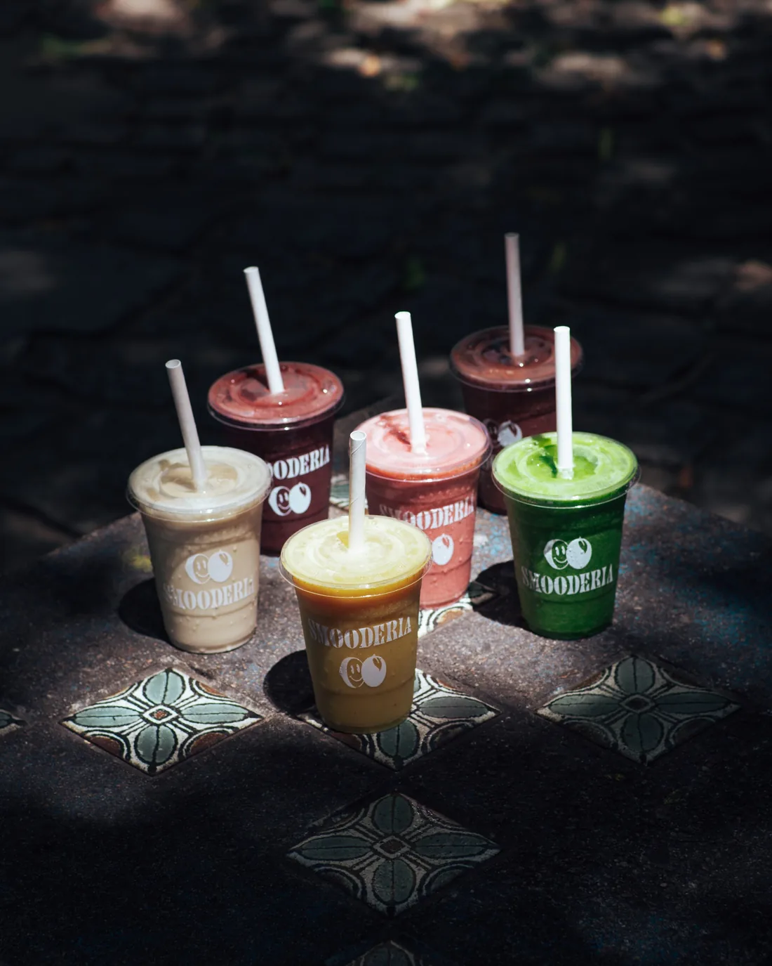 Seis copos de smoothie de cores diferentes sobre uma mesa de pedra